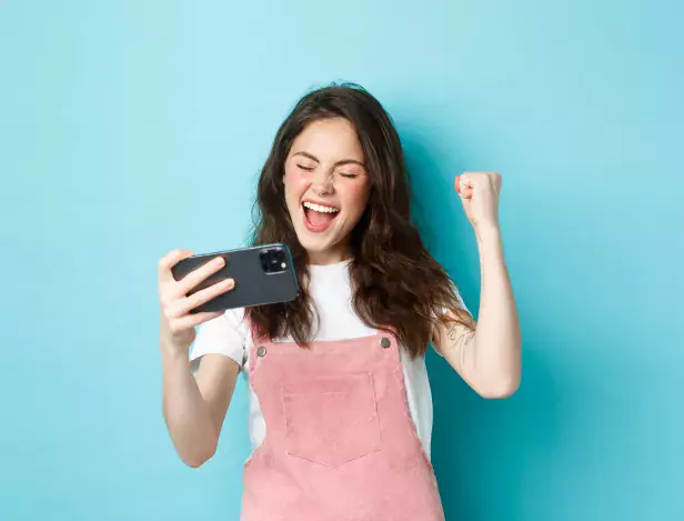 cheerful-cute-girl-winning-in-online-video-game-on-smartphone-making-fist-pump-and-shouting-yes-with-joy-standing-over-blue-background-and-triumphing